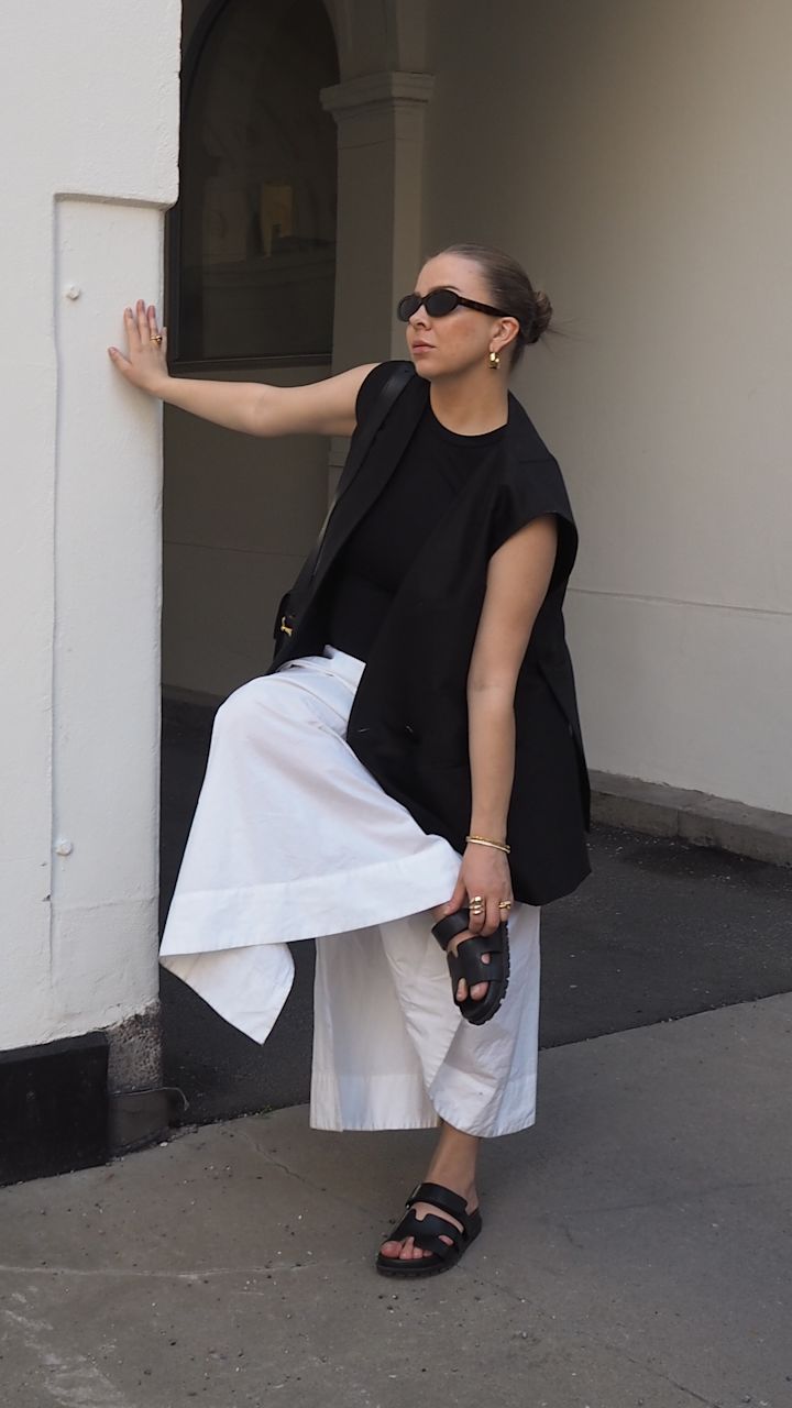 Kvinna i svart väst, svart topp, vita byxor och svarta sandaler lyfter en fot medan hon håller i en vit vägg i en urban miljö.