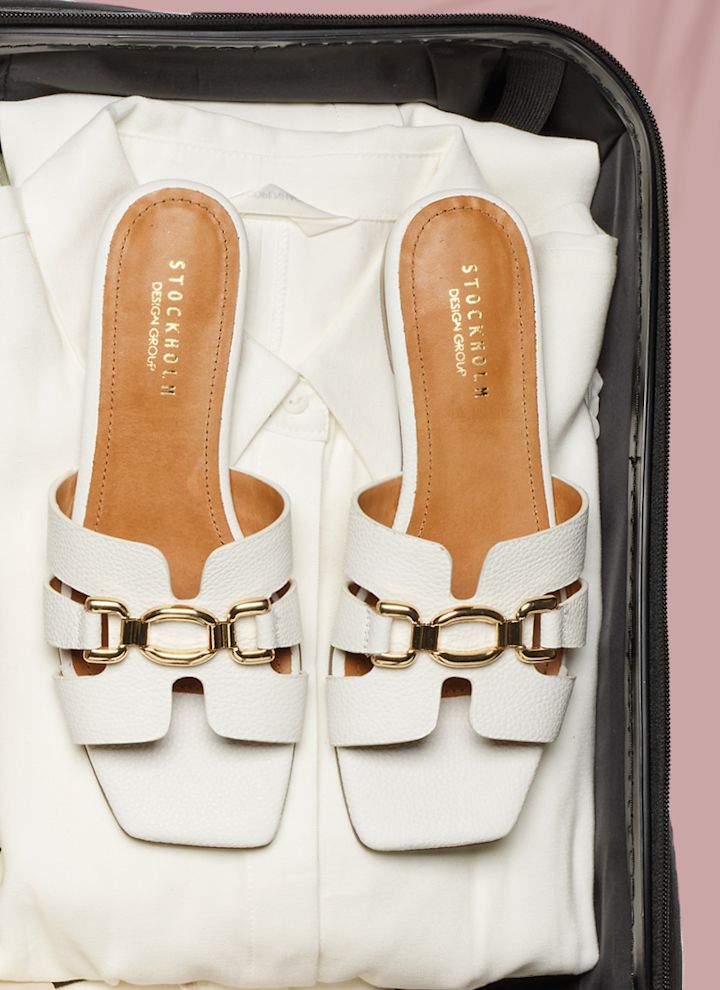 Vita platta sandaler med gulddetaljer i en packad resväska med vita kläder. Eleganta sommarsandaler med breda remmar.