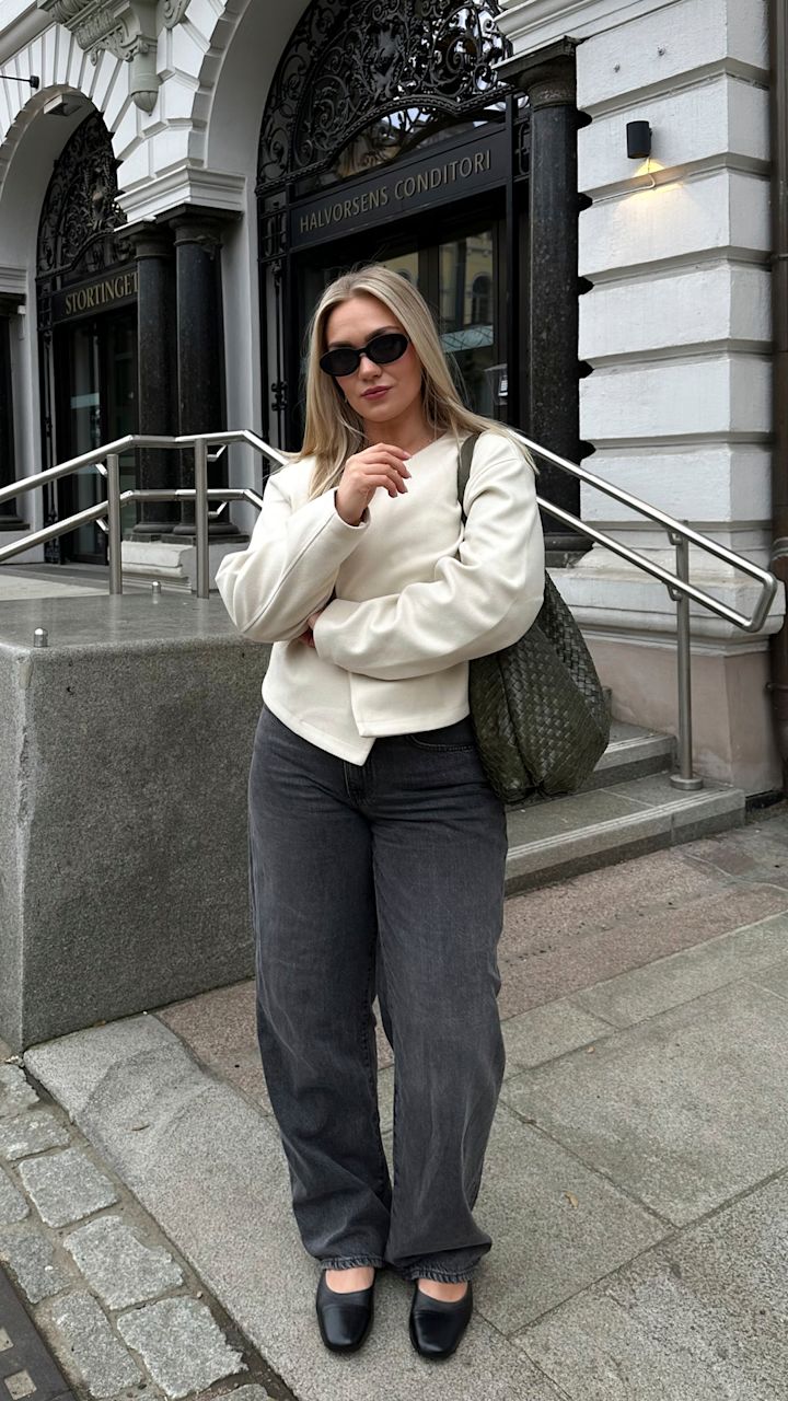 Kvinna med solglasögon, vit tröja, grå byxor och svart väska står framför en ingång med skylten "Halvorsens Conditori" i en stadsmiljö.