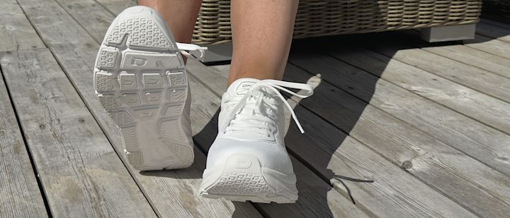 Hvite joggesko med snøring vises på en person som sitter på en treterrasse med flettet møbel i bakgrunnen.