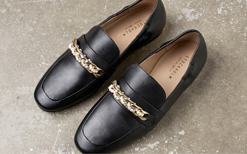 Svarta skinnloafers med dekorativt guldkedja och pärlor, placerade på en grå betongyta.