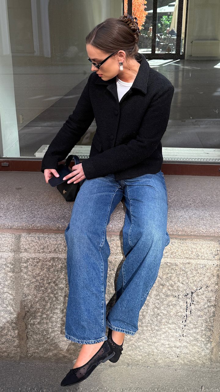 Kvinna med glasögon och svart jacka som sitter på en stenbänk, tittar ner och öppnar en liten svart väska, iklädd blå jeans och svarta skor med nät-detaljer.