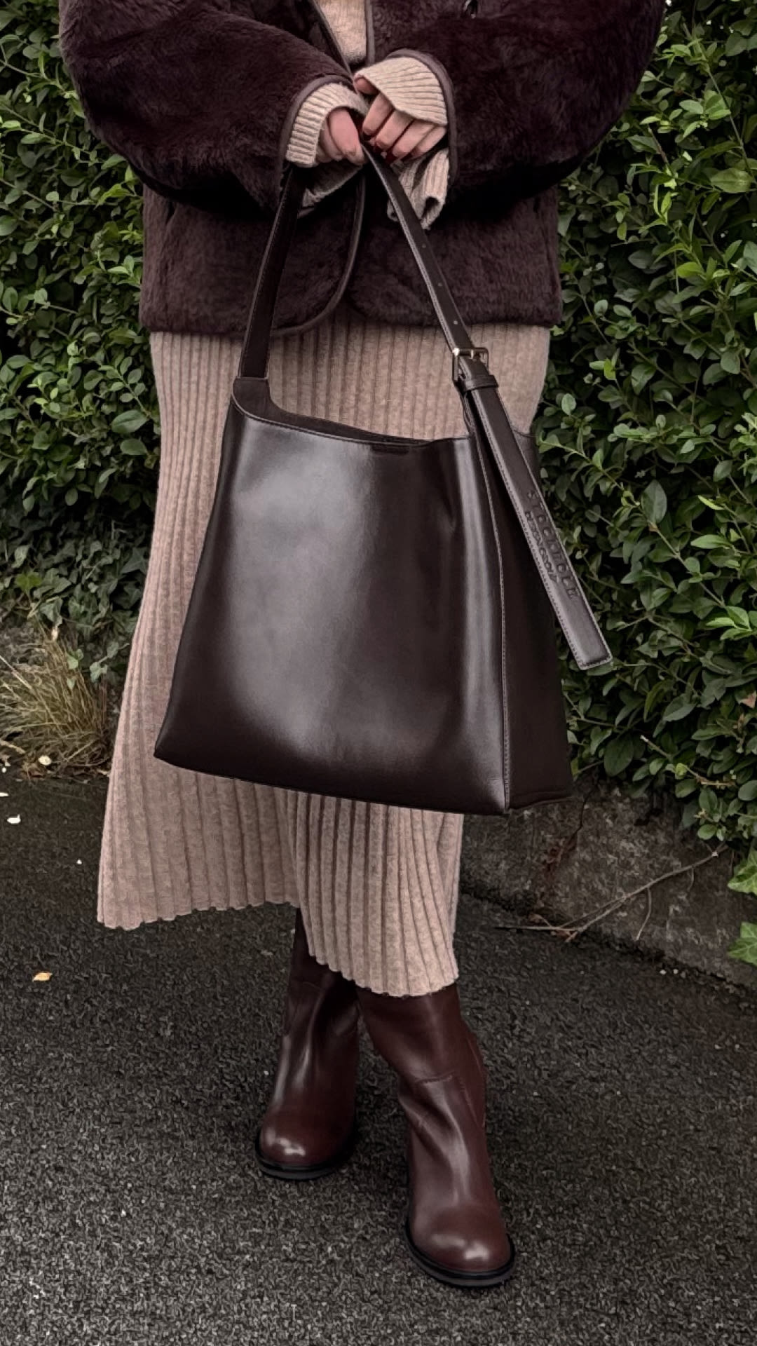 Person i brun pälsjacka och beige stickad kjol bär mörkbruna läderstövlar och håller en matchande brun läderväska.