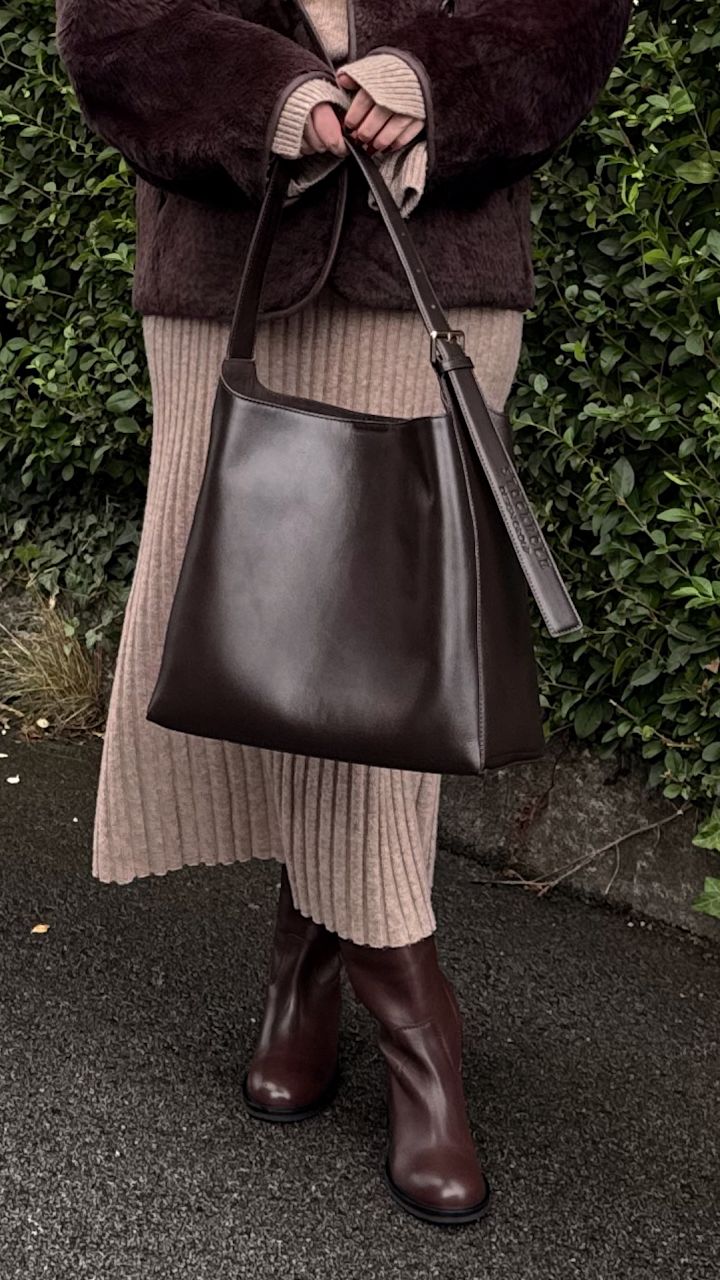 Anette Hovland i brun pelsjakke og beige strikket kjole holder mørk skinnveske, med brune høye skinnstøvler i klassisk stil fra Stockholm Design Group.