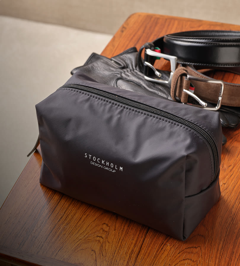 Svart toalettmappe fra Stockholm Design Group på trebord med svarte og brune skinnbelter ved siden av.