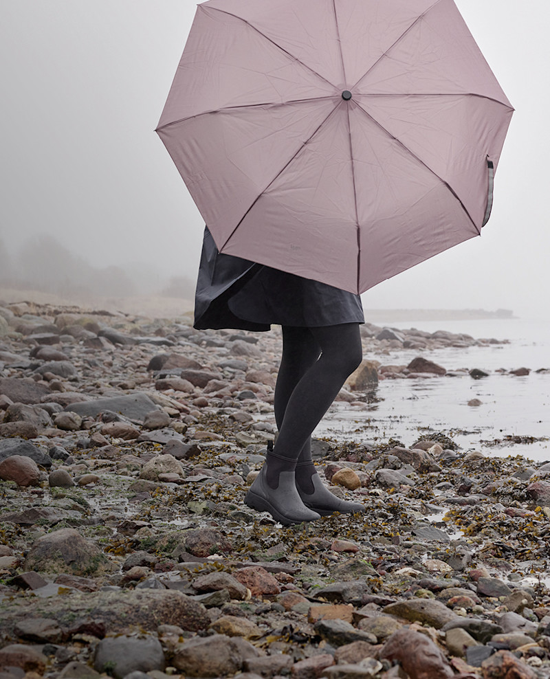 Person står på steinete strand, holder en lys paraply, iført mørke klær og støvler, med tåke i bakgrunnen.