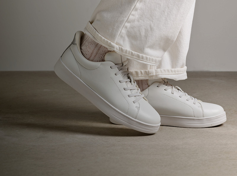 Hvite cupsole sneakers fra Stockholm Design Group med beige sokker, brukt med kremhvite bukser på en nøytral bakgrunn.