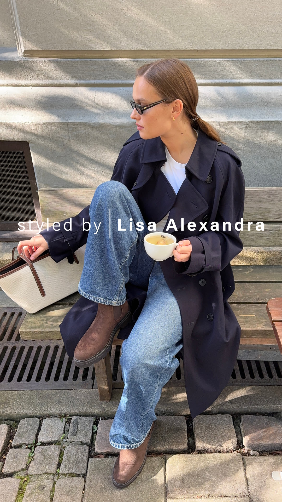 Lisa Alexandra på bänk i mörkblå kappa och jeans, bärande bruna robusta chelsea boots från Unified, håller en kaffekopp.
