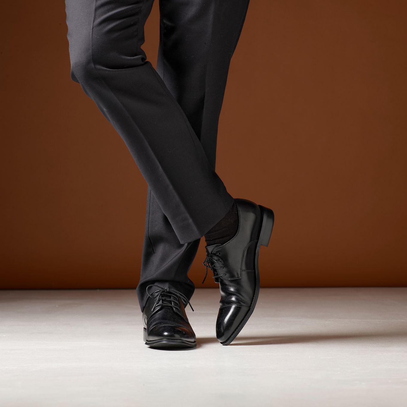 Svarte, klassiske dressko med snøring, vist med mørke dressbukser mot brun bakgrunn i en elegant, formell stil.