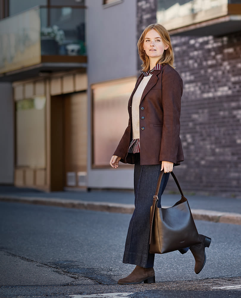 Kvinne i brun blazer og stripete bukser går på gaten med veske og brune ankelstøvletter med lav hæl i klassisk stil.