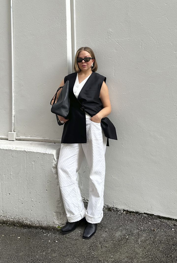 Kvinna har solglasögon och en svart väst över en vit topp, vita byxor och svarta stövlar, står mot en ljus vägg och håller en svart väska.