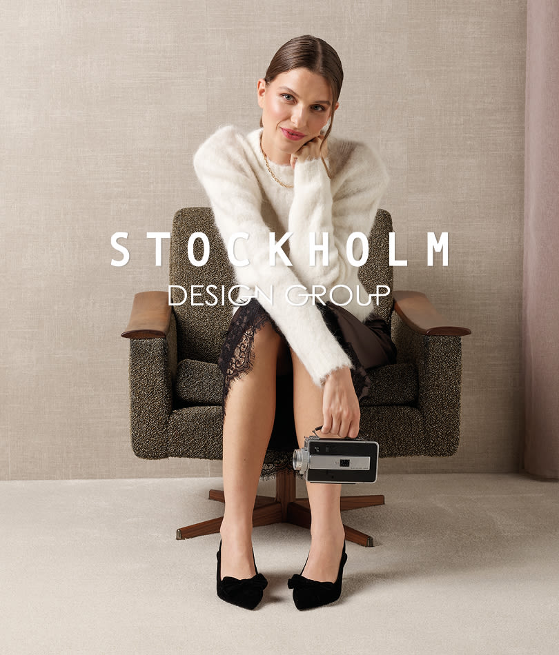Model i hvid sweater og sort nederdel sidder i lænestol med sorte elegante pumps med spids snude og Stockholm Design Group logo.
