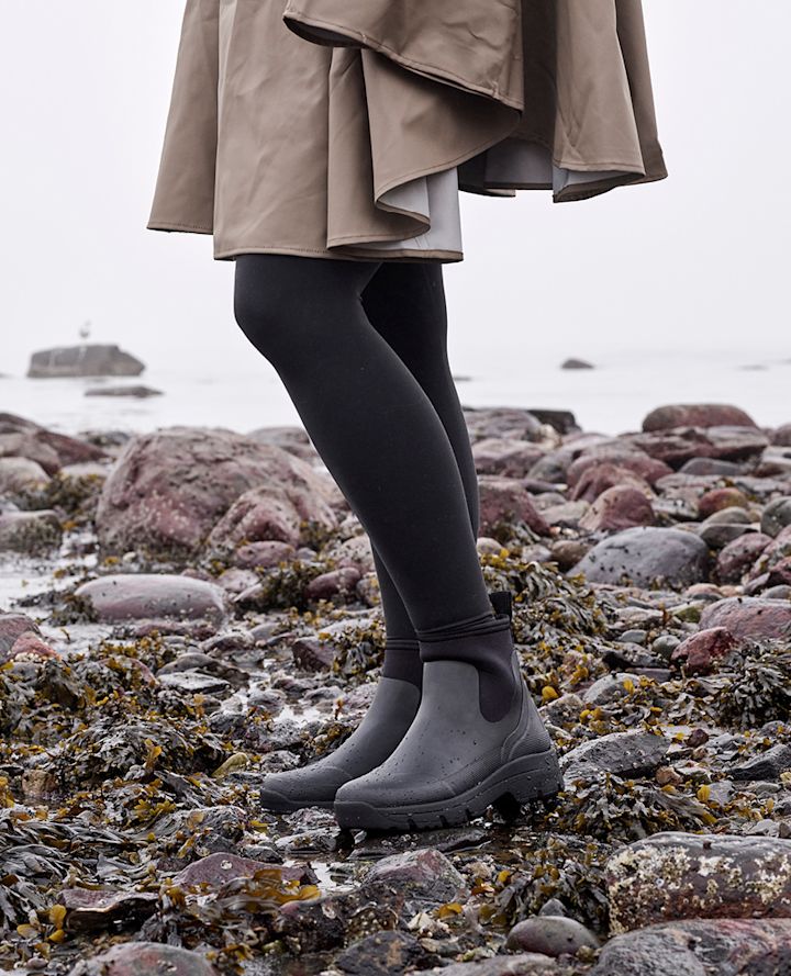 Person med svarta stövlar och svarta strumpbyxor står på en stenig strand med tång vid havet.