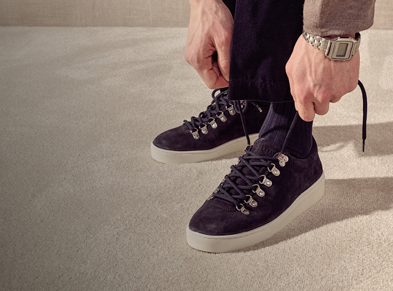 Person knytter skolisser på brune semsket sneakers fra Stockholm Design Group med hvit såle, har på seg mørke bukser og sølvklokke.