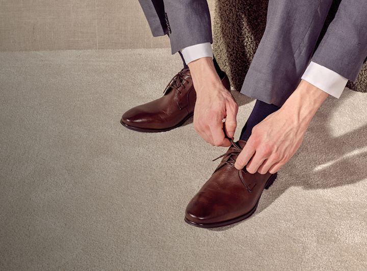 Mann i grå dress knyter snørebåndene på mørkebrune, elegante dressko fra Stockholm Design Group.