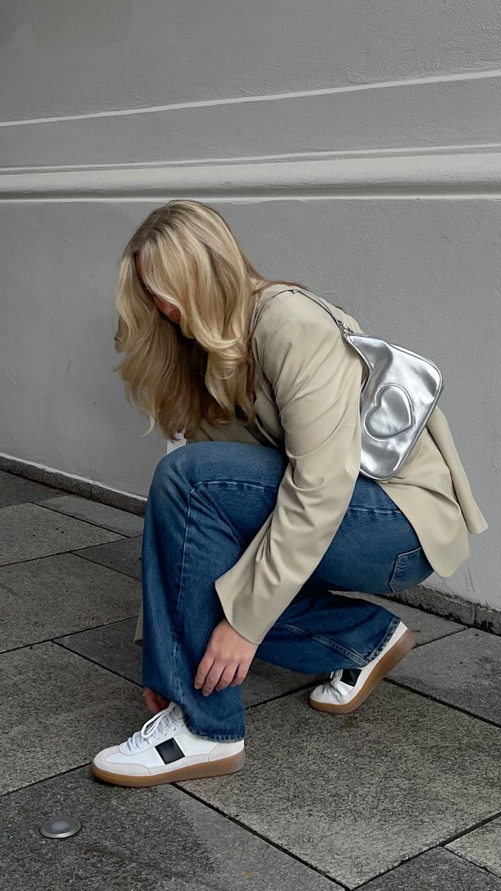 Kvinna med blont hår böjer sig ner för att knyta vita sneakers med svarta detaljer på en grå stenlagd gata, iklädd en beige blazer, blå jeans och en silverfärgad väska med hjärta.