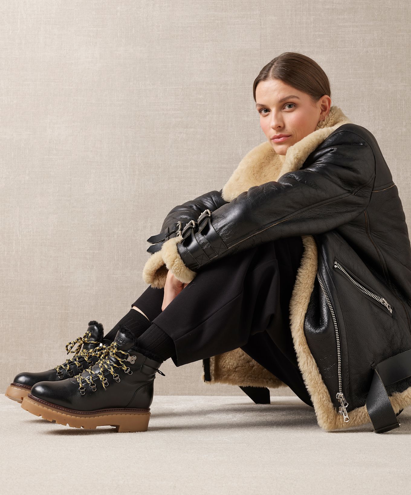 Kvinna sitter på golvet iförd en svart skinnjacka med beige foder och svarta stövlar med tjocka sulor och mönstrade skosnören.