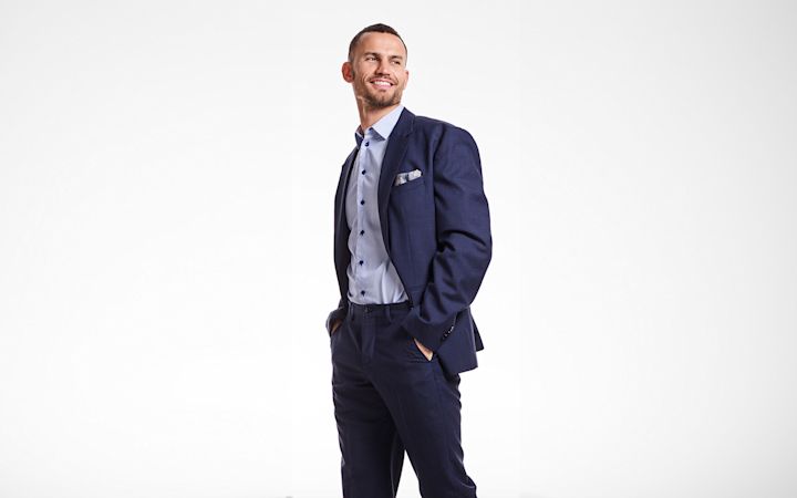 Man i mörkblå kostym med ljusblå skjorta står med händerna i fickorna och ler, mot vit bakgrund.