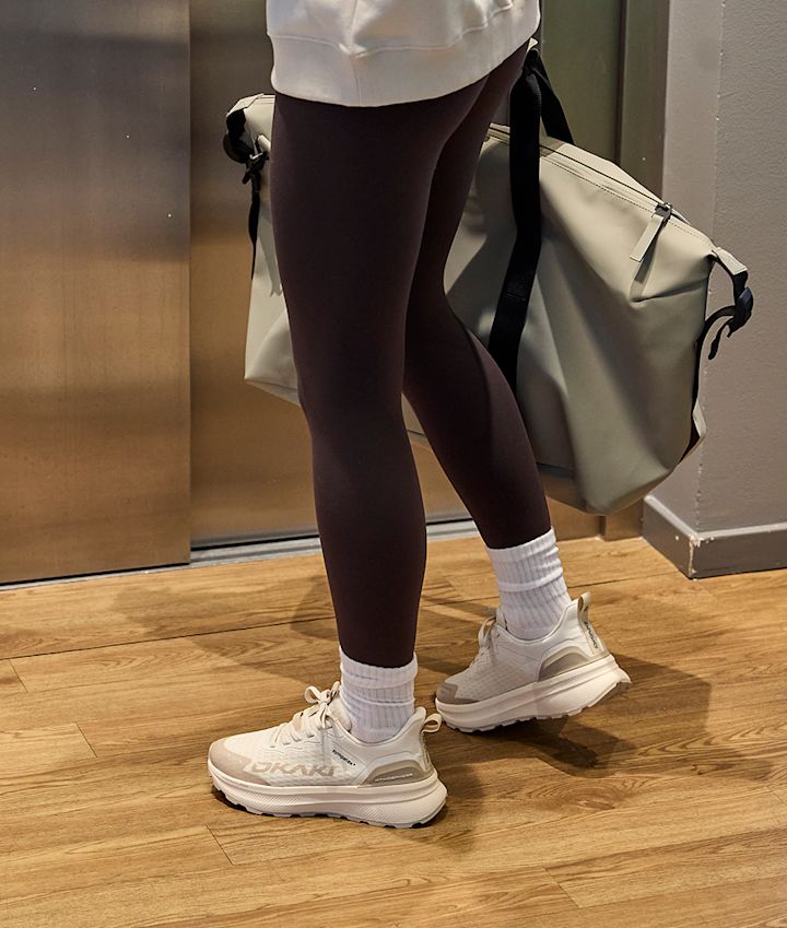 Person i bruna leggings och vita strumpor bär vita sneakers från Okaki i sportig stil, med en ljusgrå väska över axeln.