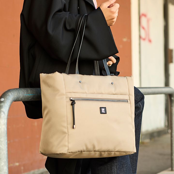 Person i svart kappa med beige tygväska med svarta handtag och dragkedja, märkt med "Unified" logotyp.