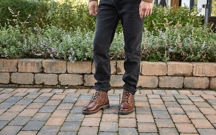 Man iklädd bruna skinnstövlar och svarta jeans står på en kullerstensbelagd gångväg med en stenmur och grön buskage i bakgrunden.
