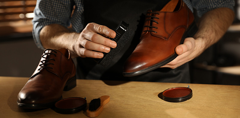 Mann pusser en brun skinnsko med en svart børste på et bord med skopussutstyr i et rom med dempet lys.