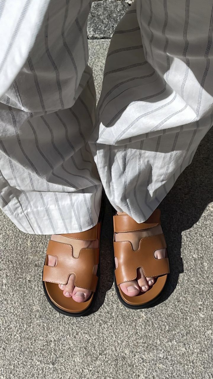 Bruna skinnsandaler med öppna tår som bärs på grått stengolv, iklädd vita byxor med tunna, mörka ränder.