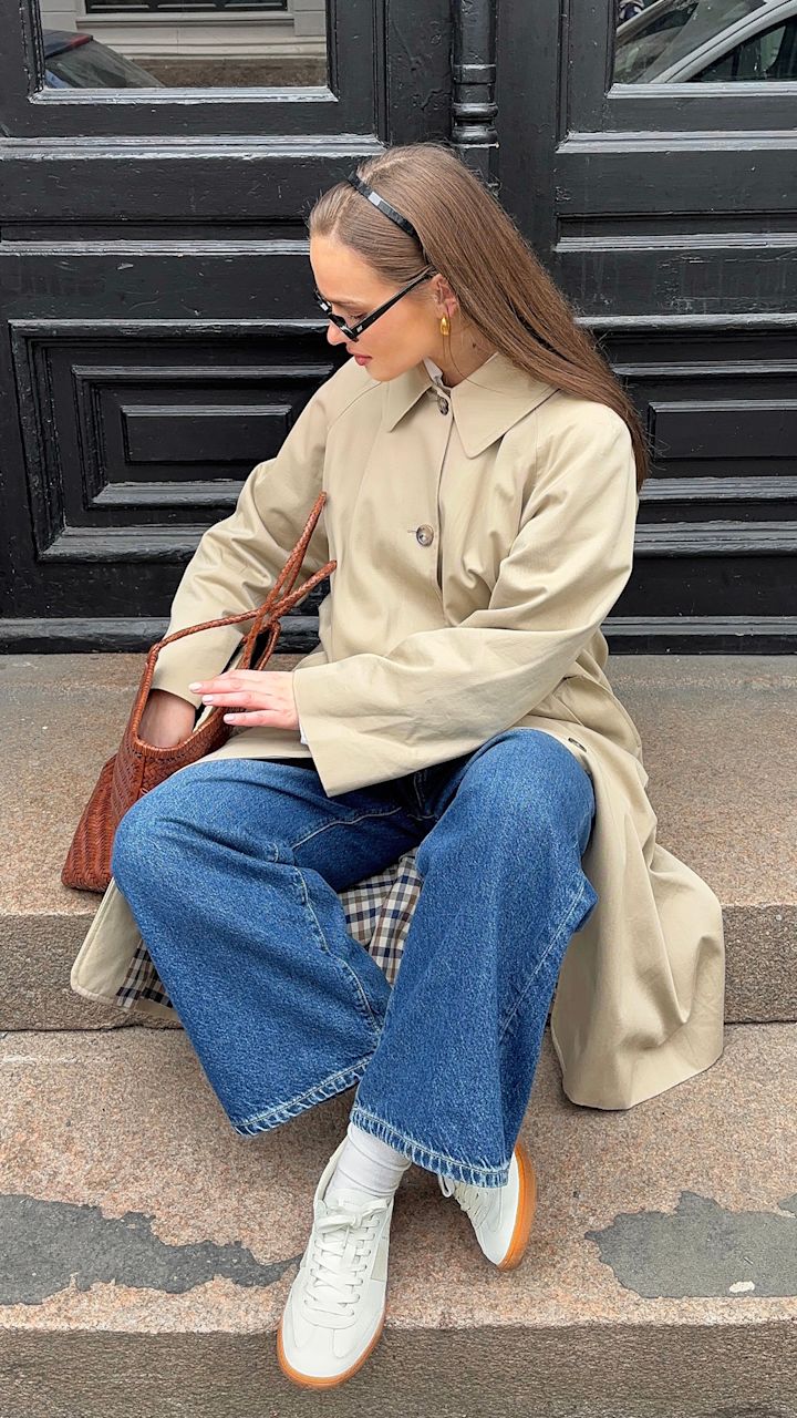 Kvinna i beige trenchcoat, blå vida byxor och vita sneakers sitter på en trappa och letar i en brun flätad väska framför en svart dörr.