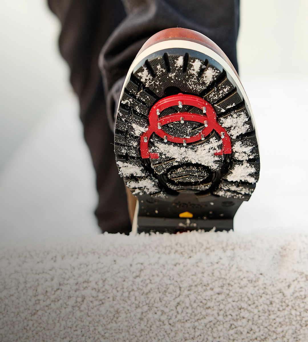 Såle av vintersko med svart mønster og rød logo, dekket med snø, på snødekt underlag.