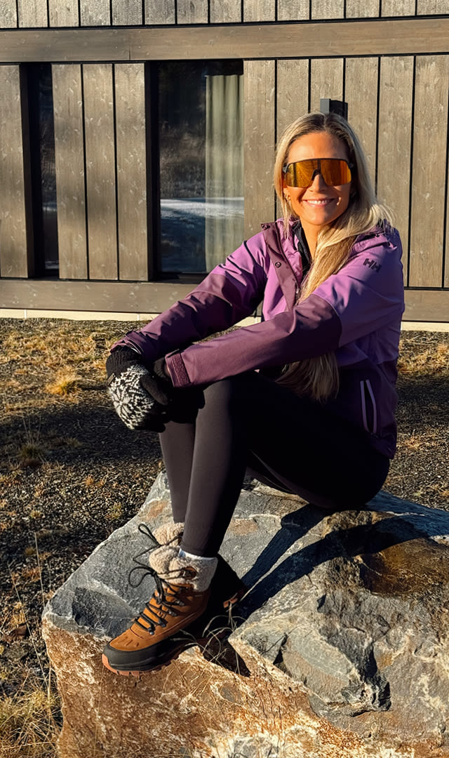 Kvinne med langt lyst hår og oransje solbriller sitter på en stor stein utenfor et moderne trehus, iført lilla jakke, svarte leggings, mønstrede votter og brune vinterstøvler.