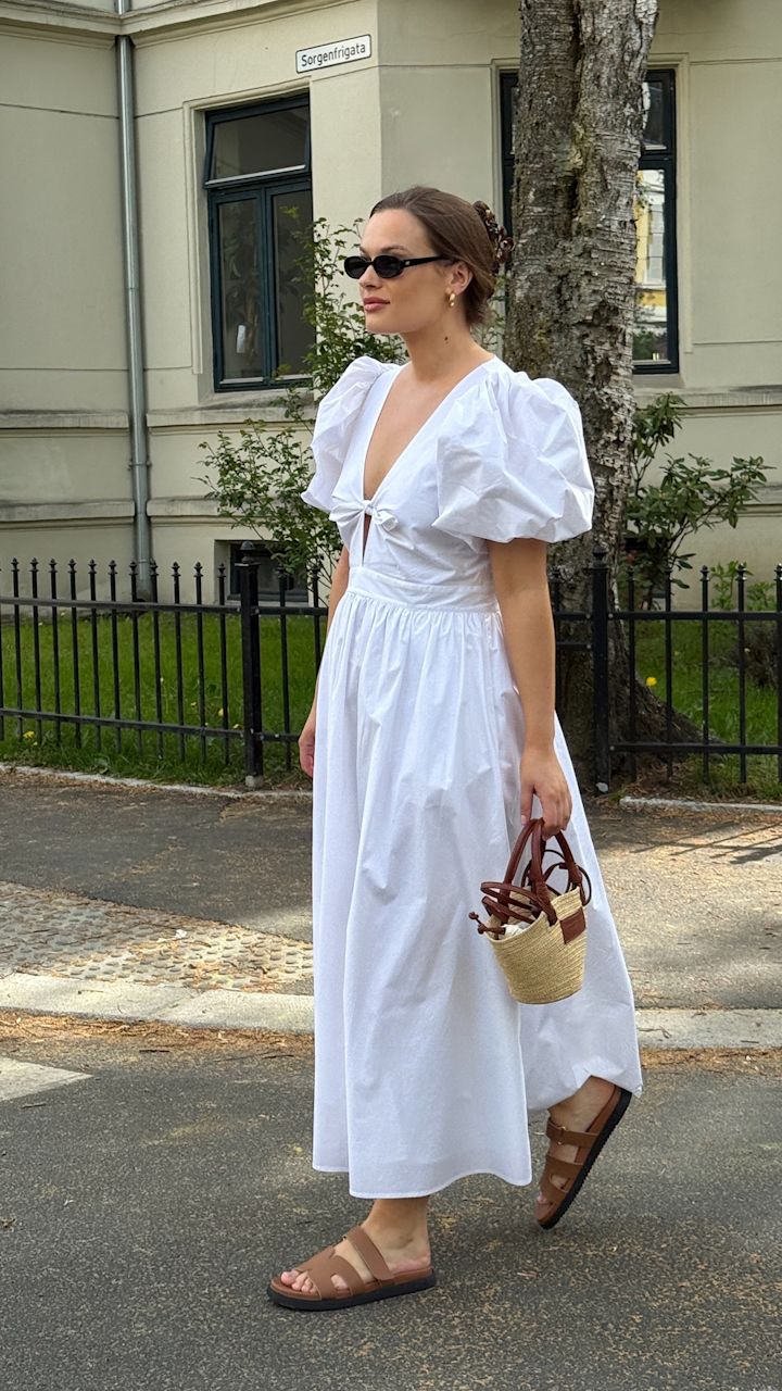 Person i vit klänning med puffärmar bär bruna lädersandaler med remmar och svart sula, håller en liten flätad väska.