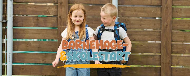 To barn med ryggsekker smiler og står foran et trestakitt med teksten "Barnehage- & skolestart" over dem.