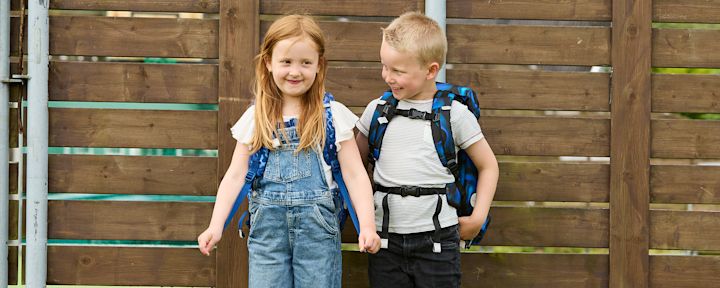 Två barn med ryggsäckar ler och står framför ett trästaket med texten "Förskola- & skolstart" ovanför dem.