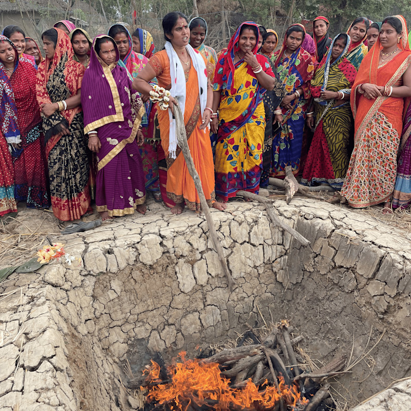 En gruppe kvinner i fargerike sarier står rundt en stor, tørr, sprukket jordgrop med en ild som brenner i bunnen, en kvinne i oransje saris rører i ilden med en lang pinne.