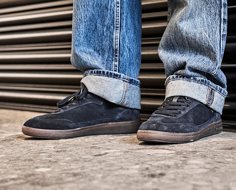 Mörkblå mocka sneakers från Unified med brun sula, burna med uppvikta blå jeans framför en metalljalusi.