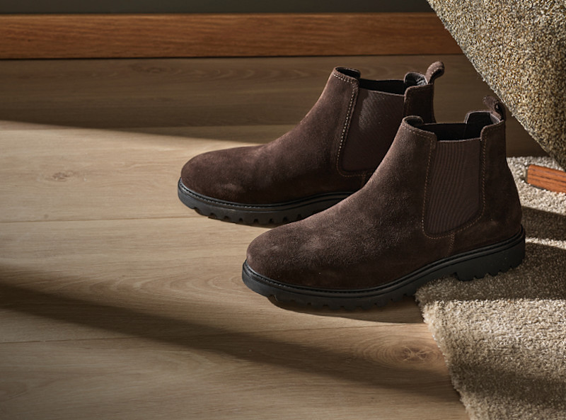 Brune chelsea-boots i semsket skinn med elastiske sideseksjoner og robust sort såle, plassert på et lyst tregulv.