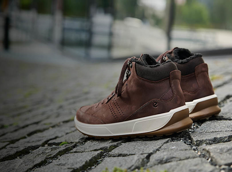 Brune høye sneakers i skinn med hvit såle og pelsfôr, plassert på brostein utendørs.
