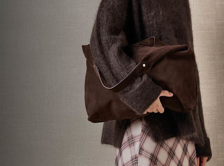 Person i mörkbrun fluffig tröja håller en brun mockaväska, med rutig kjol i beige och brunt.