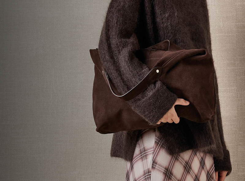 Person i mørk brun, fluffy genser holder en mørk brun semsket skinnveske fra Stockholm Design Group, med rutete skjørt under.