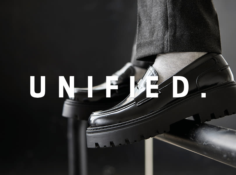 Svarta eleganta loafers med kraftig sula och texten "UNIFIED" i vitt över bilden. Bärs med grå byxor och vita strumpor.
