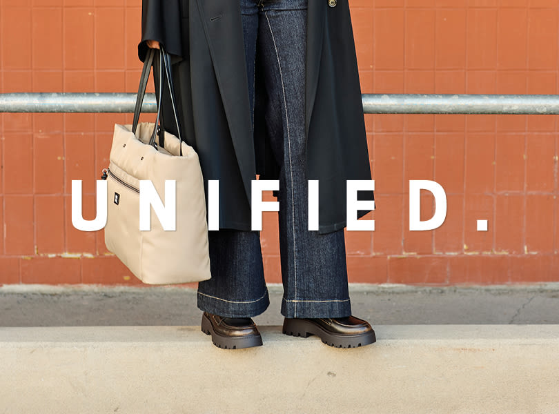 Person med svarte chunky loafers med kraftig såle, iført mørk frakk og jeans, holder en beige veske, foran oransje vegg.
