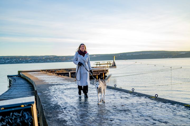 Kvinna i en ljus kappa går med hund i koppel på en isig brygga vid vattnet med berg i bakgrunden.