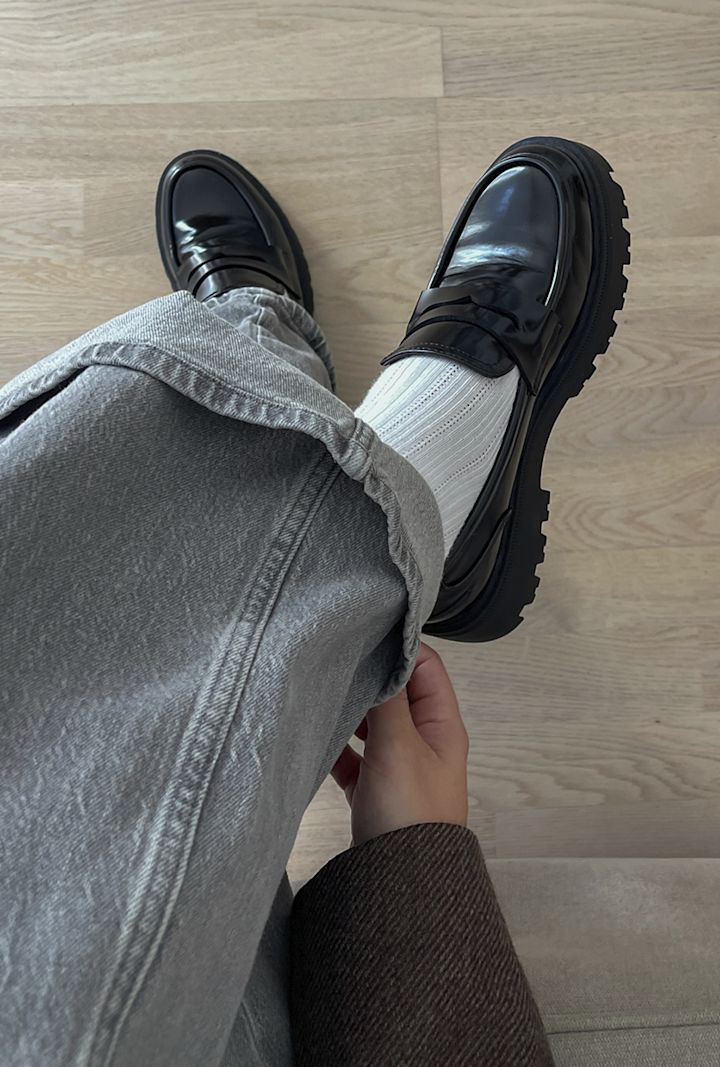 Sittande person som justerar uppvikta grå byxor över vita strumpor och svarta lackskor på ljust trägolv.