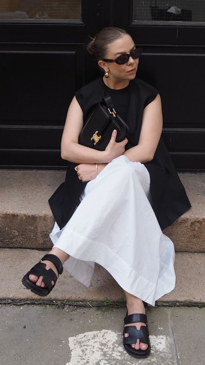 Kvinna bär svarta solglasögon, svart väst och en vit lång kjol, sitter på en trappa och håller en svart väska med guldfärgad spänne.