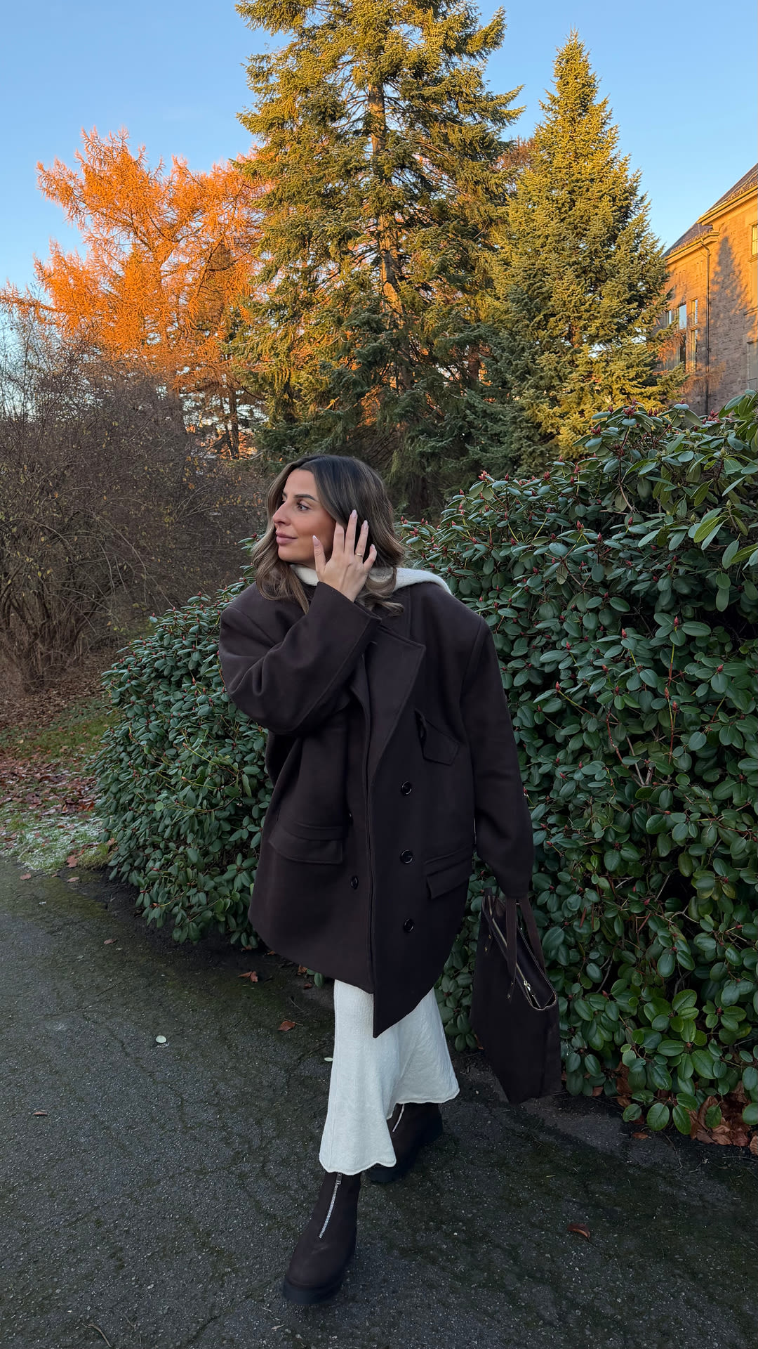 Influenser Sara Boudaya i mørk frakk og hvit kjole med brune boots fra Stockholm Design Group står foran høstfarget landskap med grønne og oransje trær.