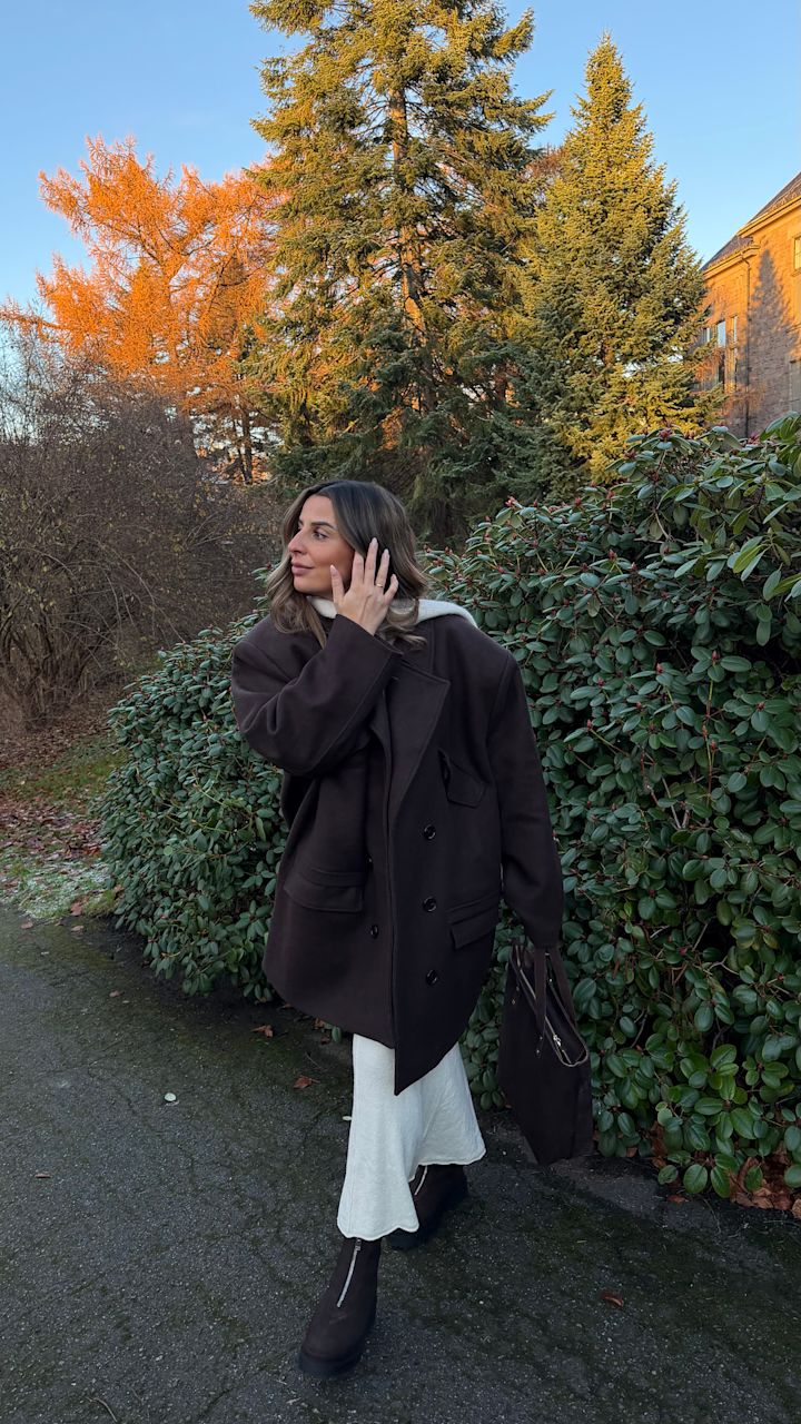 Influenser Sara Boudaya i mørk frakk og hvit kjole med brune boots fra Stockholm Design Group står foran høstfarget landskap med grønne og oransje trær.