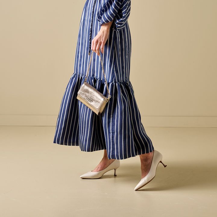 Kvinne går iført blå og hvitstripet lang kjole med volang nederst, hvite pumps og holder en liten gullfarget veske med kjedestropp.