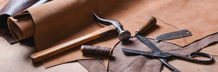 Rulle med läder, hammare, trådrullar, skräddarsax och läderverktyg placerade på ett bord med läderbitar.