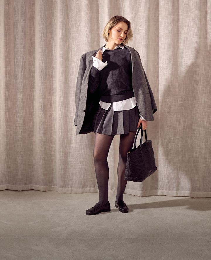 Kvinna i skoluniform med grå kavaj, veckad kjol och svarta klassiska loafers, håller en svart handväska mot beige gardin.