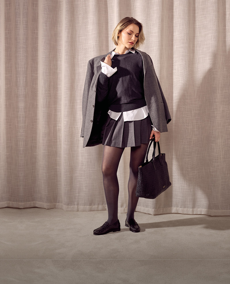 Kvinna i skoluniform med grå kavaj, veckad kjol och svarta klassiska loafers, håller en svart handväska mot beige gardin.